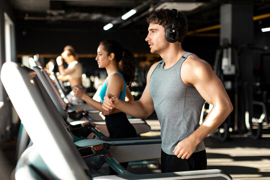 Young European muscular man running on treadmill and listening music, doing cardio workout in modern gym
