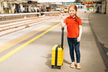 Young girl waiting at a train station with a yellow suitcase and a hopeful expression on a sunny day