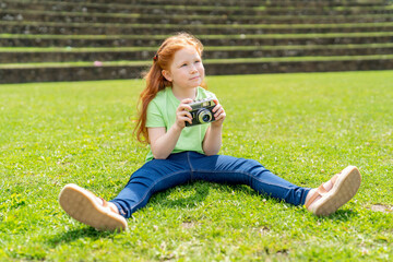 Fototapeta premium Young girl sits on grassy field with vintage camera in hand, enjoying sunny day in outdoor setting