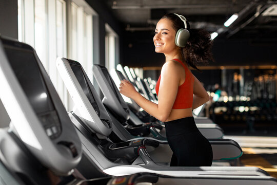 Beautiful happy fit woman running on treadmill and enjoying music wearing headphone during sports training in modern gym