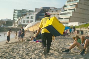 unrecognizable street ice cream vendor carrying a cooler on a sandy beach near modern buildings