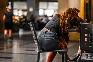 Young Woman Sitting Alone at a Table with Her Head Resting, Deep in Thought or Exhaustion, in a...