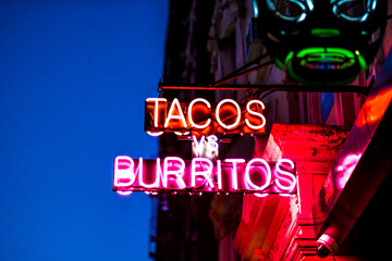Vibrant Neon Sign Featuring a Playful "Tacos vs Burritos" Theme Glows Brightly Against a Deep Blue Night Sky in an Urban Setting