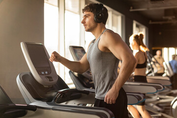 Handsome man listening music in headphones while jogging on treadmill at gym, working out during...