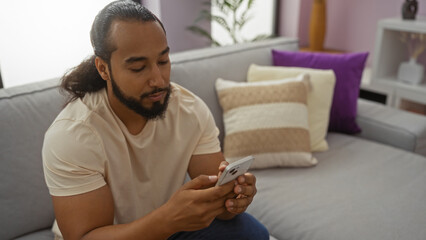 Obraz premium Young man with beard using smartphone indoors in a modern living room with cozy decor, suggesting a relaxed atmosphere, and highlighting home technology use.