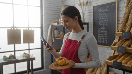 Young woman in a bakery wearing a red apron holding a pastry while looking at her phone in a cozy interior