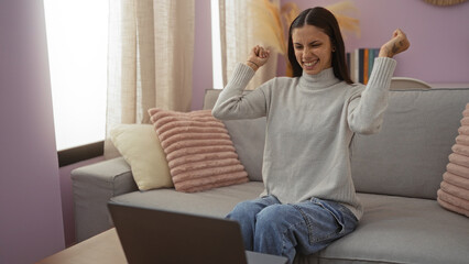 Happy young hispanic woman celebrating indoors in an apartment living room with a laptop