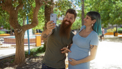Man and woman enjoying a videoconference call in an urban park, pointing to her pregnant belly, celebrating pregnancy together outdoors on a sunny day