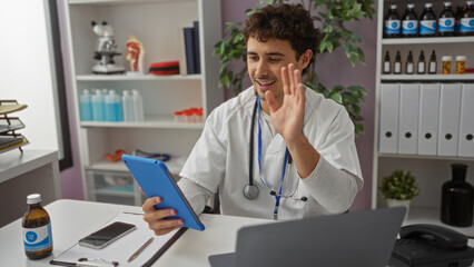 Young hispanic man in clinic smiles while using tablet for a video call, showcasing a modern healthcare environment with digital devices and medical equipment.