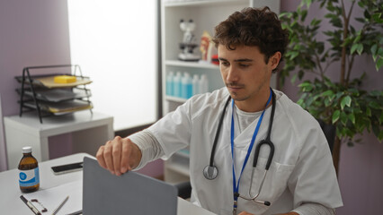 Young man in a clinic setting working at a desk with a stethoscope, surrounded by medical equipment...