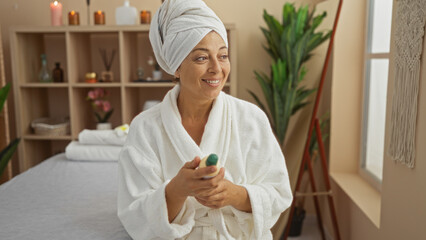 Woman relaxing in a spa, wearing a bathrobe and towel, holding a bottle of lotion, with candles and plants in the background, smiling in a serene setting.