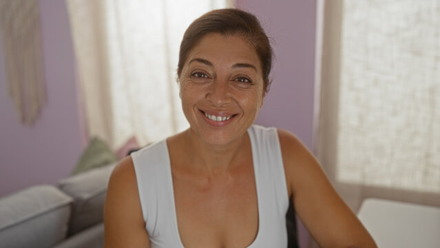 Hispanic woman smiling in living room, showing joyful expression with brunette hair, seated indoors, wearing white top, in warm home environment with purple walls.