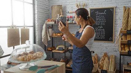 Hispanic woman making a video call inside a bakery, surrounded by bread and pastries, wearing an apron, against a backdrop of a chalkboard menu and paper bags hanging from a window.