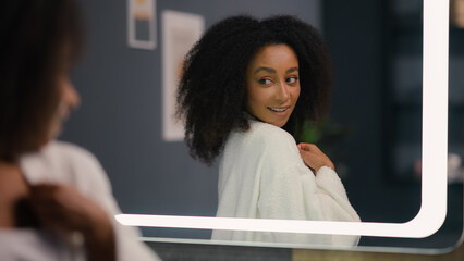 Happy smiling satisfied African American woman looking in mirror feeling joyful pampering in morning bathrobe smile touching face and curly hair girl female skin care haircare cosmetics getting ready