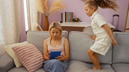 Obraz premium Woman using tablet while daughter jumps on sofa in cozy living room, illustrating family life and motherhood dynamics indoors.