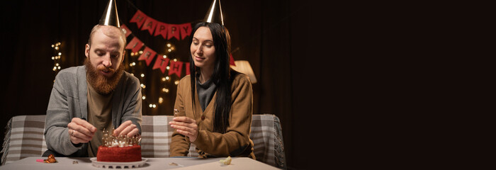Young couple in party hats sitting near table and man lightning candles on birthday cake, celebrating together at home. family enjoying domestic anniversary party. Copy space