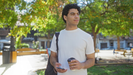 Fototapeta premium Handsome young hispanic man standing outdoors in a city park with coffee and phone in hand, looking thoughtful.