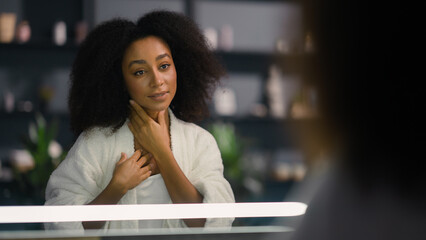 African American happy smiling healthy woman looking at mirror reflection enjoy perfect skin tone after facial mask cream girl pampering touch hair spa procedure at home natural beauty morning routine