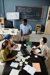 Vertical shot of young African American teacher standing at desk talking to students explaining...