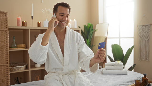 Middle-aged hispanic man in a spa wellness center talking on mobile while holding a credit card looking content in a robe