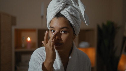 A beautiful young woman with a towel on her head and wearing a bathrobe applies skincare product to...