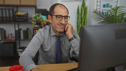 A mature hispanic man is working in an office, smiling while looking at his computer screen with a red mug on his desk and bookshelves in the background.