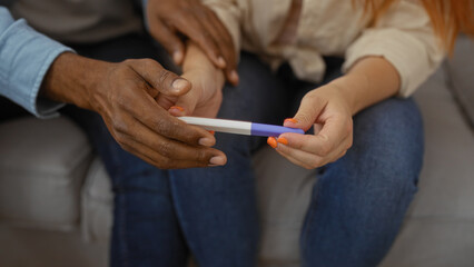Interracial couple holding pregnancy test in cozy living room, suggesting excitement and bonding in their home.