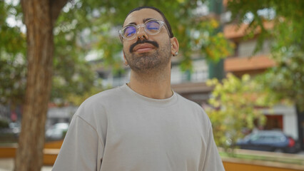 Young hispanic man with glasses and moustache stands outdoors in an urban park setting with trees...