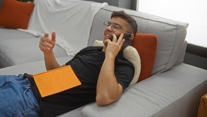 Young man relaxing indoors on sofa in modern living room with phone and book enjoying conversation at home