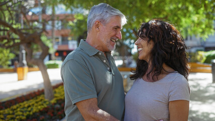 Man and woman smiling lovingly at each other in a sunny urban park, surrounded by greenery, showcasing a middle-aged couple in a joyful, romantic relationship outdoors.