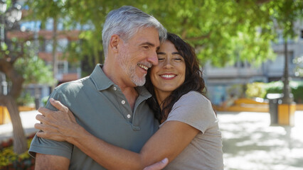 Fototapeta premium Middle-aged couple embracing warmly in a sunny city park, exuding love and happiness, with a backdrop of urban greenery and buildings.