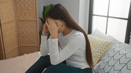 Young woman sitting indoors on bed appearing stressed in bedroom setting with light coming through...