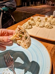 A hand holding a bite-sized khinkali, with the rest of the dumplings neatly arranged on a plate nearby. The rustic presentation and natural lighting highlight the authenticity and freshness
