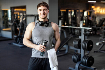 Sporty young man standing in gym after workout, holding water bottle and towel after training, guy...