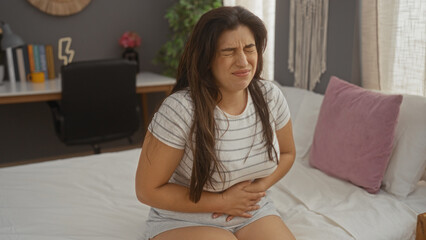 Woman in casual clothing clutching her stomach in pain while sitting on bed in a cozy living room with a blurred desk and decorations in the background