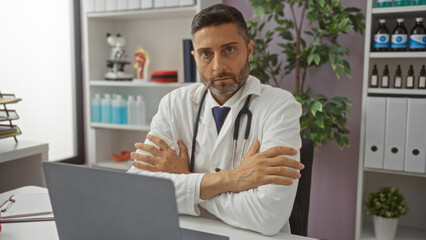 Fototapeta premium Doctor seated at a desk indoors in a modern clinic with arms crossed looking focused in a white coat with a stethoscope on a bright day with folders and bottles around.