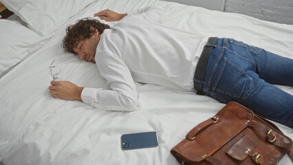 Young man sleeping on bed in office attire with phone and leather bag nearby indicating work-life balance or exhaustion in a contemporary interior setting