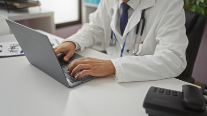Doctor working on laptop in clinic room wearing white coat using modern technology in healthcare setting, hands typing on keyboard in professional environment.