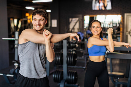 Fit European couple, man and woman, doing arms stretching while warming up before workout in modern gym interior