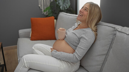 Pregnant woman with blonde hair relaxing on a couch in a cozy living room, enjoying a peaceful moment at home with a warm drink, highlighting the beauty of pregnancy and anticipation.