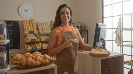Young woman working in a bakery shop standing behind the counter with a confident smile in a cozy indoor setting filled with fresh bread and pastries