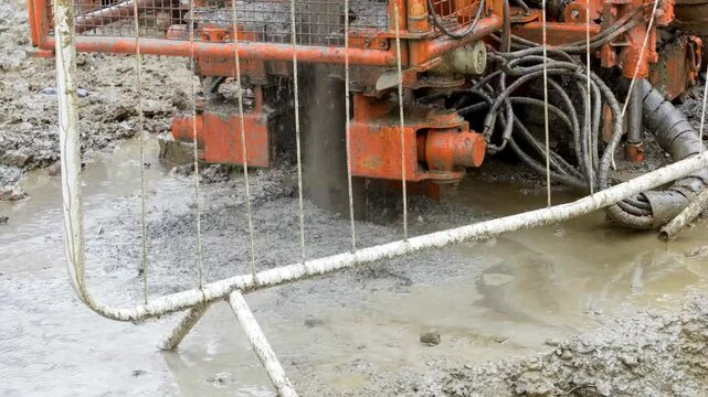 Construction site shows piling rig drilling equipment operating in muddy conditions during a rainy day
