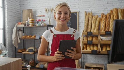 Young woman baker with short blonde hair holding a tablet in a well-lit bakery shop surrounded by bread and pastries