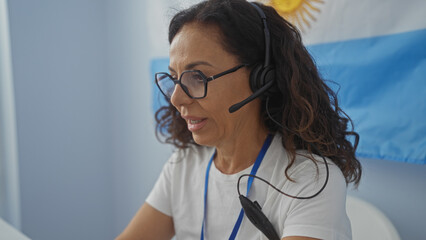 Woman wearing glasses and a headset in a room with an argentinian flag, focusing on work indoors during an electoral process.