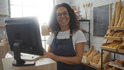 Woman working happily at a bakery counter wearing an apron and glasses, surrounded by various types...