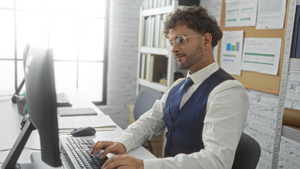 Fototapeta premium Handsome hispanic man focused on computer work in modern office setting with natural light illuminating workspace, embodying professionalism and dedication in urban environment.