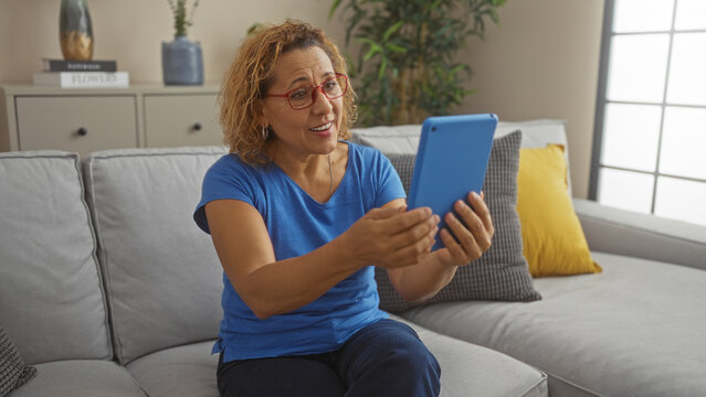 A mature hispanic woman with curly hair is sitting on a sofa in a living room, happily holding a blue tablet, wearing glasses and a blue shirt.