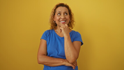 A beautiful middle-aged hispanic woman with curly hair smiles thoughtfully while standing against a...