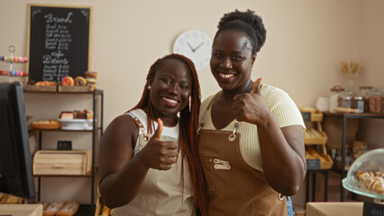 African american women bakers smiling and giving thumbs up in a cozy bakery shop interior with bread and pastries display.