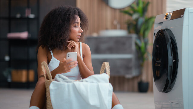 African American woman ethnic girl housewife do housework housekeeping basket with cloth dirty clothes reading instruction using washing machine laundry appliance chatting mobile phone smartphone hold